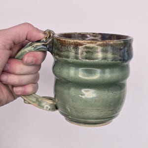 May include: A handmade ceramic mug with a green glaze and a brown rim. The mug has a unique, textured design and a large handle. The mug is held in a hand. This stoneware mug is perfect for coffee or tea.
