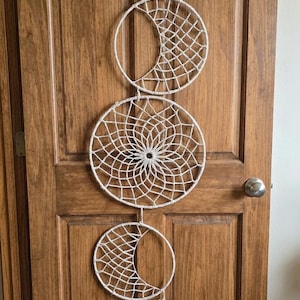 May include: A white macrame wall hanging featuring a series of circular and crescent moon shapes. The dream catcher is suspended against a wooden door. The design includes intricate woven patterns within the circular frames, creating a bohemian aesthetic.