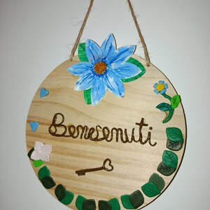 Può includere: Un cartello di benvenuto rotondo in legno con un fiore blu, foglie verdi e la parola "Benvenuti" in corsivo marrone. Il cartello è decorato con una chiave e cuori, ed è sospeso da una corda di iuta.