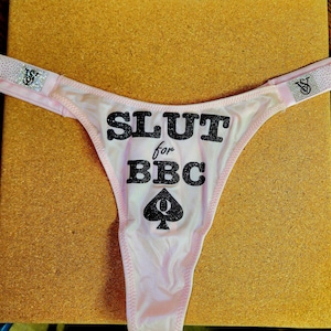 Könnte beinhalten: Hellrosa String-Tanga-Slip mit dem Text "SLUT for BBC" in schwarzem Glitzer. Unter dem Text befindet sich das Pik-Symbol. Die Träger haben ein Strass-Design und das "VS"-Logo.