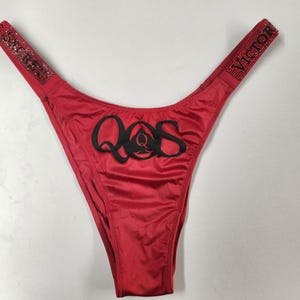 Könnte beinhalten: Roter String-Tanga mit schwarzem "QQS"-Text auf der Vorderseite. Die Träger tragen das Wort "VICTORIA" in Strasssteinen. Der Stoff ist glänzend und glatt.