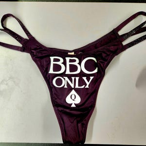 May include: Plum-colored thong underwear with the text "BBC ONLY" in white, along with a white spade symbol. The straps have decorative embellishments.