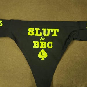 May include: Black thong underwear with the text "SLUT for BBC" in bright yellow. The underwear has a spade symbol and the text "SIZE M" on the side straps. The word "TERS" is also visible on the straps.