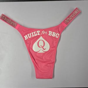 May include: Bright pink thong underwear featuring rhinestone-encrusted side straps with the text "VICTORIA SECRET". The front displays a white graphic of a spade symbol with the letter "Q" inside, and the text "BUILT for BBC" printed above.