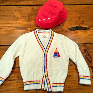 May include: A cream-colored knit cardigan with a textured checkerboard pattern, featuring a V-neck, button-down front, and red, yellow, and blue striped trim. An embroidered sailboat patch is on the chest. A matching red cap with three white buttons.