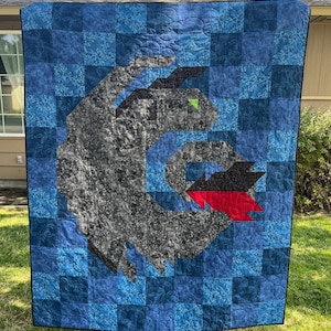 May include: A handmade quilt featuring a stylized dragon design. The quilt showcases a patchwork of blue squares with a central dragon motif in gray, black, and red. The dragon's eye is a small green triangle.