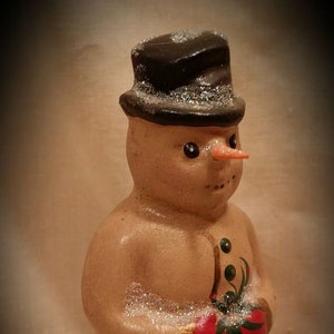 May include: A small, hand-painted snowman figurine with a black top hat, a carrot nose, and green and red holly painted on its body.