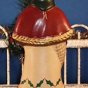 Puede incluir: Una figura navide&ntilde;a vintage, que presenta una figura con un gorro verde y una capa roja. La figura tiene un cuerpo color crema con detalles de acebo y dorado. La base es verde con un dise&ntilde;o de tronco de &aacute;rbol marr&oacute;n.