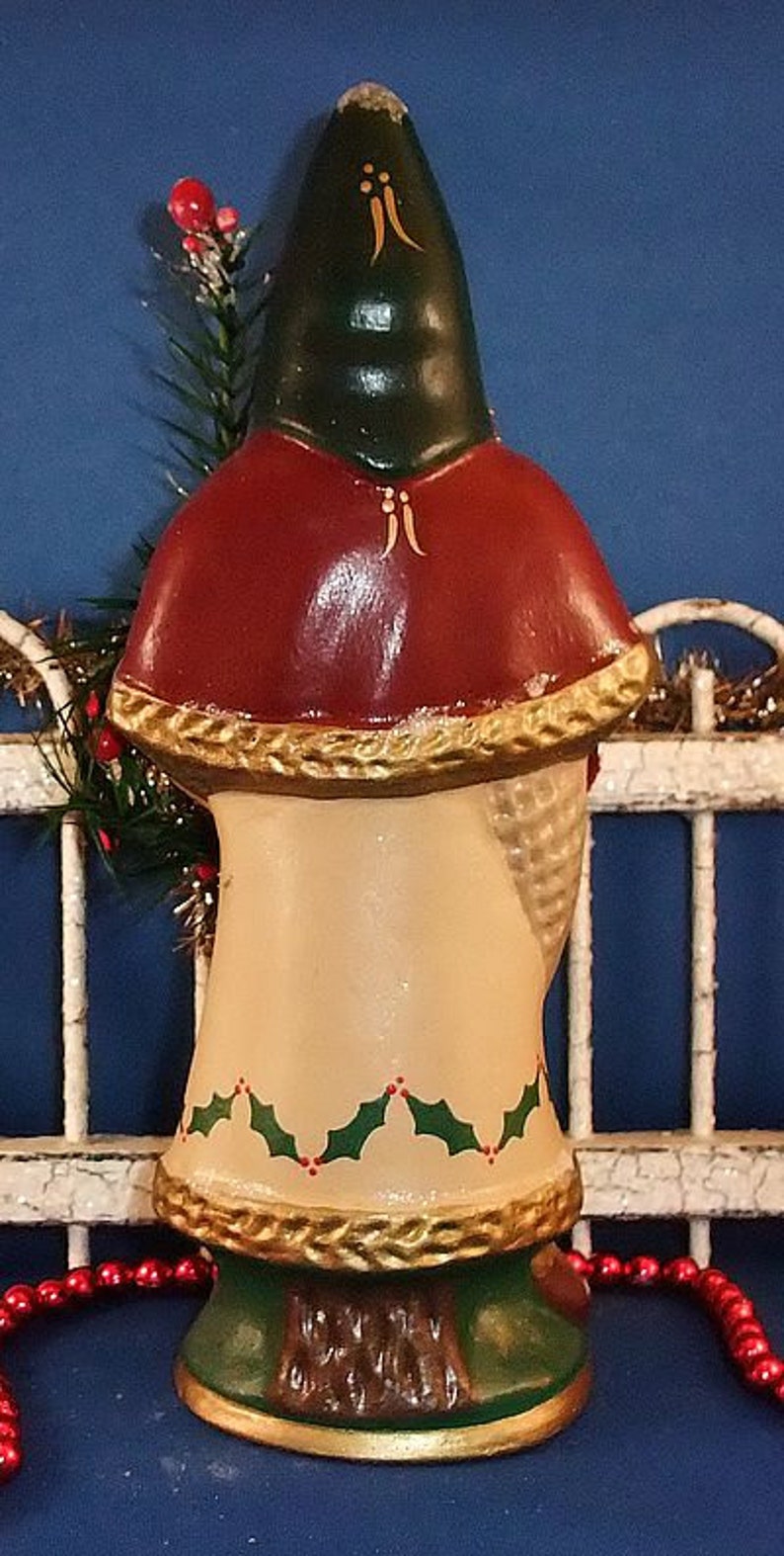 Puede incluir: Una figura navide&ntilde;a vintage, que presenta una figura con un gorro verde y una capa roja. La figura tiene un cuerpo color crema con detalles de acebo y dorado. La base es verde con un dise&ntilde;o de tronco de &aacute;rbol marr&oacute;n.