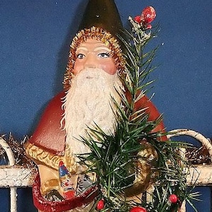Puede incluir: Una figura vintage de Pap&aacute; Noel con barba blanca, abrigo rojo y gorro verde. La figura sostiene un peque&ntilde;o &aacute;rbol de Navidad y un adorno dorado. La figura de Pap&aacute; Noel est&aacute; decorada con oropel dorado y bayas rojas, evocando un tema festivo.
