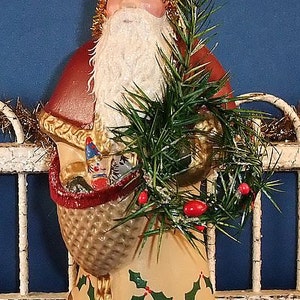 Puede incluir: Figura vintage de Pap&aacute; Noel con gorro verde, barba blanca y abrigo rojo. La figura sostiene una cesta con regalos y una ramita de vegetaci&oacute;n con bayas rojas. La base es verde con detalles dorados y el dise&ntilde;o es festivo.