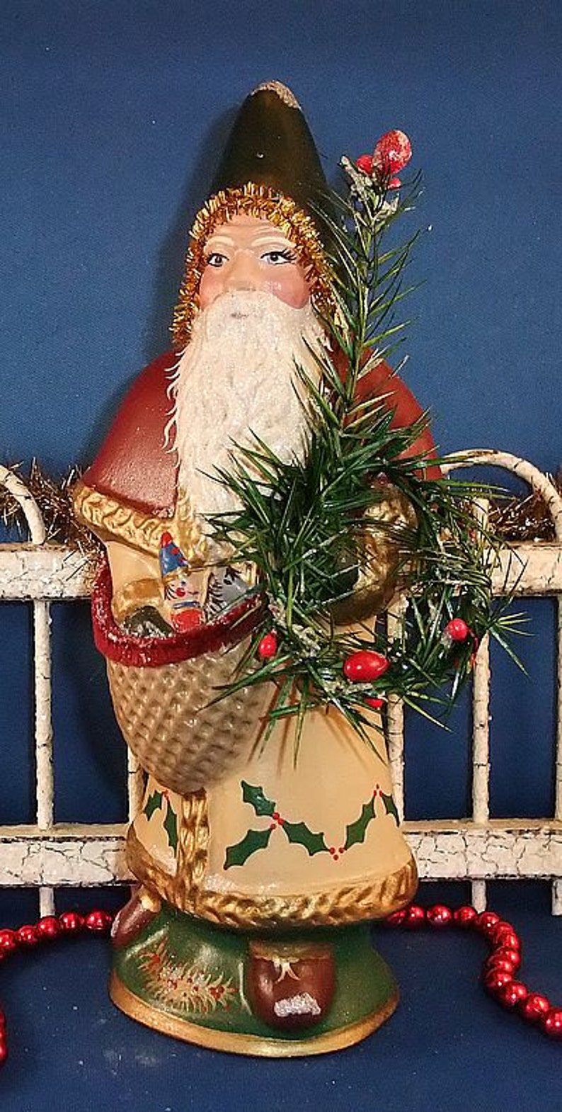 Puede incluir: Figura vintage de Pap&aacute; Noel con gorro verde, barba blanca y abrigo rojo. La figura sostiene una cesta con regalos y una ramita de vegetaci&oacute;n con bayas rojas. La base es verde con detalles dorados y el dise&ntilde;o es festivo.