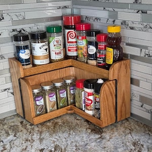 May include: A wooden corner spice rack with two tiers, holding various spice jars with black lids and labels. The rack is made of light-colored wood and is positioned on a countertop against a tiled backsplash.