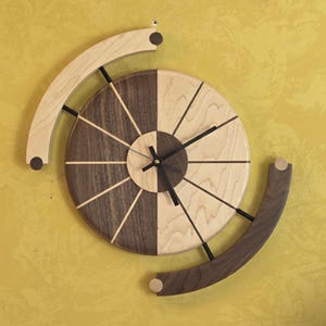 Könnte beinhalten: Eine zweifarbige Wanduhr aus Holz mit einem Spiraldesign. Das Zifferblatt ist in helle und dunkle Holzsegmente unterteilt. Zwei gebogene Holzstücke erstrecken sich von der Uhr und tragen zu ihrer einzigartigen Ästhetik bei. Die Zeiger der Uhr sind schwarz.