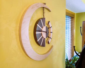 May include: A wooden wall clock with a crescent moon shape and a circular clock face. The clock face features a radial design with alternating dark and light wood segments. The clock is suspended by a rope and hangs on a yellow wall.