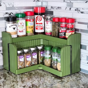 May include: A green, two-tiered corner spice rack filled with various spice jars. The jars have red, green, and black lids, and labels with text like "Kinder," "Italian," and "Lawry's." The rack is made of wood and sits on a countertop.