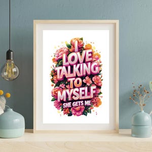 Può includere: Una stampa artistica incorniciata con il testo "I LOVE TALKING TO MYSELF SHE GETS ME" in rosa e bianco, circondata da fiori rosa, arancioni e gialli. L'opera d'arte è esposta su una superficie bianca.