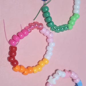 Könnte beinhalten: Drei kreisförmige Armbänder aus bunten Kunststoffperlen. Ein Armband zeigt einen Regenbogenverlauf, ein weiteres besteht aus blauen, weißen und grünen Tönen, und das dritte enthält hellblaue, rosa und weiße sternförmige Perlen.