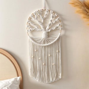 May include: Cream macrame wall hanging with a tree design. The tree is made from knotted rope within a circular wooden frame. Below the tree, long rope strands hang, decorated with small wooden beads, creating a bohemian style, ideal for home decor.