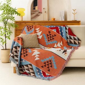 Puede incluir: Una manta tejida con un patrón geométrico en tonos naranja, azul, marrón y blanco cubre un sofá blanco. Una almohada beige descansa sobre el sofá. La manta tiene un borde con flecos y un diseño del suroeste.