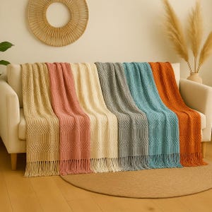 Puede incluir: Una colección de seis mantas decorativas texturizadas en varios colores, incluyendo beige, rosa, crema, gris, turquesa y naranja. Cada manta tiene flecos en el borde inferior y está colocada sobre un sofá blanco. Las mantas tienen un patrón tejido.