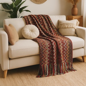 May include: A cream sofa draped with a colourful chevron-patterned throw. The blanket has red, green, and gold tones with a fringed edge. Beige and tan cushions add to the cosy living room setting. The sofa has wooden legs.