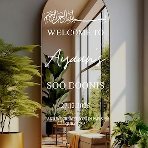 May include: A large, arched mirror with a gold frame. The mirror has white text that reads "WELCOME TO Ayaan's SOO DOONIS 27.12.2025" and a quote from the Quran. The mirror reflects a room with plants and a couch.