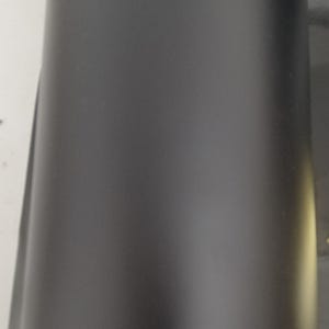 May include: A close-up view of a matte black cylindrical object. The surface is smooth and reflects minimal light, creating a sleek, modern appearance. The object's shape suggests it could be a container or part of a larger product.