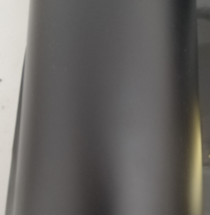 May include: A close-up view of a matte black cylindrical object. The surface is smooth and reflects minimal light, creating a sleek, modern appearance. The object's shape suggests it could be a container or part of a larger product.
