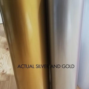 May include: Close-up of two rolls of shiny vinyl material, one gold and one silver. The text "ACTUAL SILVER AND GOLD" is printed in black across the bottom of the image. The vinyl has a smooth, reflective surface.