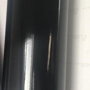 May include: A roll of glossy black vinyl material. The surface reflects light, creating highlights and shadows. The material is tightly rolled, showing a smooth, shiny finish. The background includes a white sheet with printed text.