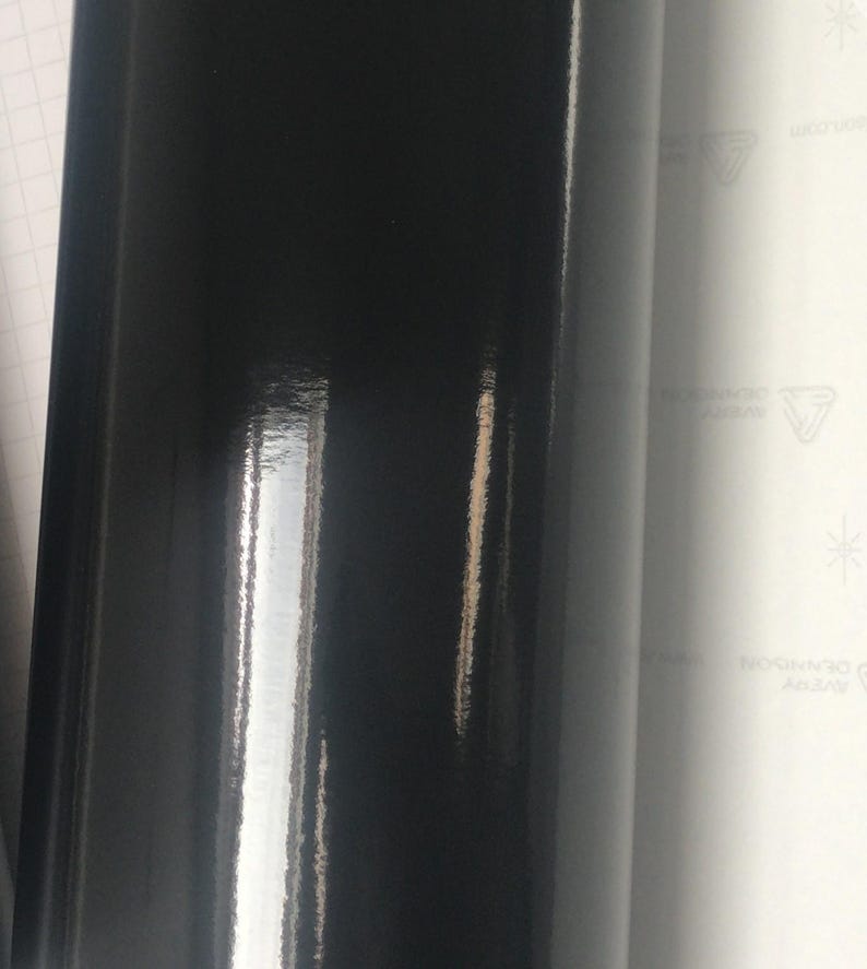 May include: A roll of glossy black vinyl material. The surface reflects light, creating highlights and shadows. The material is tightly rolled, showing a smooth, shiny finish. The background includes a white sheet with printed text.