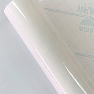 May include: A roll of glossy, off-white vinyl film. The film is tightly rolled, revealing a smooth, reflective surface. The backing paper is visible, featuring a grid pattern and printed text.