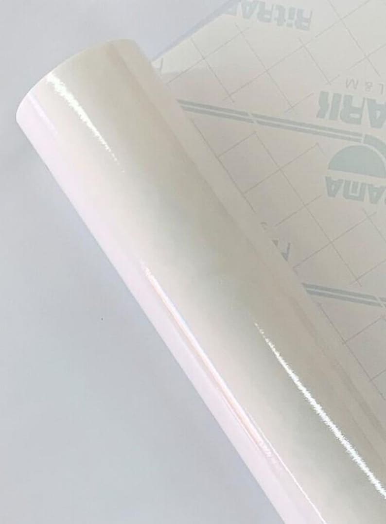 May include: A roll of glossy, off-white vinyl film. The film is tightly rolled, revealing a smooth, reflective surface. The backing paper is visible, featuring a grid pattern and printed text.