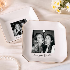 Puede incluir: Un letrero de madera rústico blanco con la palabra "Welcome" escrita a mano. El letrero tiene un aspecto envejecido y un elemento floral decorativo. El fondo es una imagen borrosa de una casa.