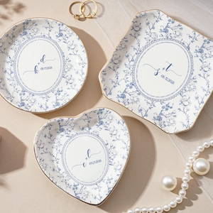 May include: Three ceramic ring dishes in round, square, and heart shapes. Each dish has a white background with blue floral patterns and gold trim. The dishes have personalized text in the centre.
