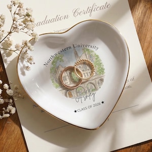 May include: A heart-shaped white ceramic trinket dish with a gold rim, featuring a Northwestern University illustration and the name "Ashley" with "CLASS OF 2026". Two gold rings rest inside the dish. A graduation certificate is in the background.