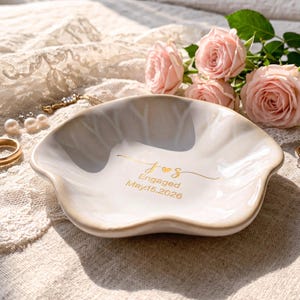 May include: A white ceramic trinket dish with a scalloped edge and a beige rim. The center features gold lettering that reads J and S with a heart icon, followed by the text Engaged May 15, 2026. The dish is surrounded by light pink roses.