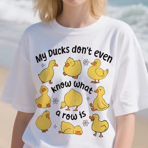 May include: White t-shirt with a cartoon duck design and the phrase "My Ducks don't even know what a row is". The design includes yellow ducks in different poses, small flowers, and stars. A fun, casual graphic tee.