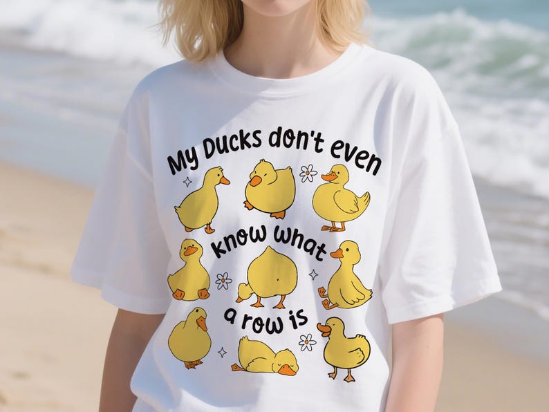 May include: White t-shirt with a cartoon duck design and the phrase "My Ducks don't even know what a row is". The design includes yellow ducks in different poses, small flowers, and stars. A fun, casual graphic tee.