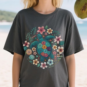 May include: Dark gray t-shirt featuring a teal turtle with a floral design. The turtle's shell is adorned with colorful flowers, surrounded by a wreath of leaves and more flowers in pink, yellow, and blue. The shirt is a casual style.