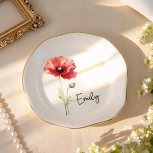 Può includere: Un piatto in ceramica bianca con bordo dorato, con un design di papavero ad acquerello. Il piatto ha una forma irregolare e il nome "Emily" è scritto sopra. Una cornice dorata, una collana di perle e fiori sono sullo sfondo.
