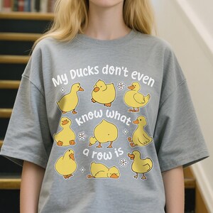 May include: A grey t-shirt with the text "My Ducks don't even know what a row is" and illustrations of yellow cartoon ducks. The shirt is a casual, comfortable style.