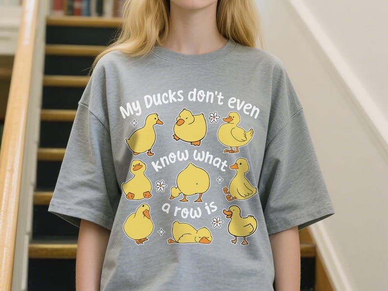 May include: A grey t-shirt with the text "My Ducks don't even know what a row is" and illustrations of yellow cartoon ducks. The shirt is a casual, comfortable style.