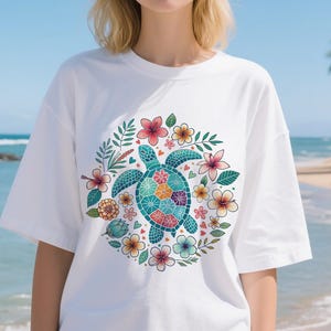 May include: White t-shirt featuring a colorful sea turtle design surrounded by flowers and leaves. The turtle has a mosaic-like pattern on its shell, with a variety of colors. The flowers are in shades of pink, orange, and yellow.