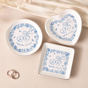 Pode incluir: Conjunto de três pratos de cerâmica branca para anéis em forma redonda, de coração e quadrada. Cada prato apresenta um design floral azul com dois corações conectados, iniciais e uma data. Bordas douradas.