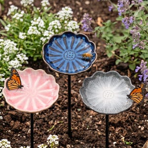 May include: Three flower-shaped garden stakes with shallow basins in blue, pink, and grey. The blue basin features a small bee, while the pink and grey basins each feature a monarch butterfly. Each basin is mounted on a thin black metal rod.