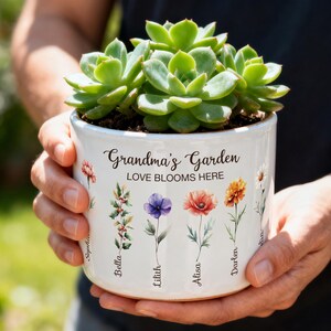 Op de afbeelding: Een witte keramische bloempot met de tekst "Grandma's Garden LOVE BLOOMS HERE" en bloemenillustraties. De pot bevat een groene vetplant. De namen Sophia, Bella, Lilith, Alisa en Darten staan op de pot.
