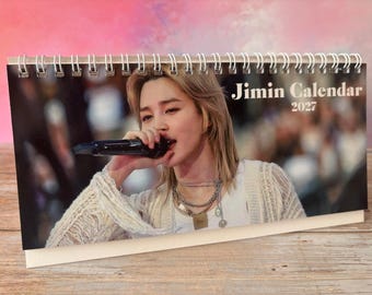 Könnte beinhalten: Ein spiralgebundener Tischkalender mit einem Foto einer Person mit blondem Haar, die in ein schwarzes Mikrofon singt. Die Person trägt ein weißes Oberteil, eine strukturierte cremefarbene Strickjacke und silberne Ketten. Text: Jimin Calendar 2027.