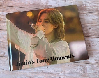 Könnte beinhalten: Ein rechteckiges Fotobuch mit dem Titel Jimin's Tour Moments, das auf dem Cover eine Person mit hellbraunem Haar in einem weißen langärmeligen Hemd zeigt, die während eines Auftritts auf einer Bühne mit warmer Beleuchtung ein Mikrofon hält.